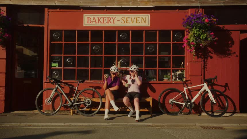 Two female cyclists enjoy a relaxing break on a bench in front of a red bakery on a sunny day