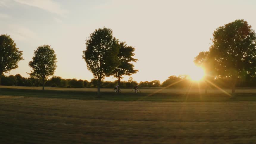 A scenic tracking shot follows two silhouetted cyclists riding their bikes through the countryside during a beautiful golden hour sunset
