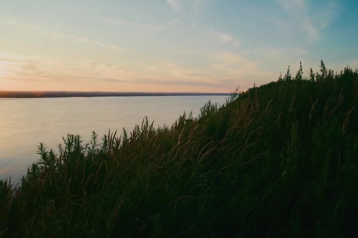 A tranquil sunset over a wide river seen from a silhouetted grassy hill