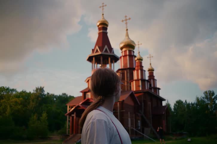 A woman from behind gazes up at a beautiful wooden Orthodox church with golden domes at dusk