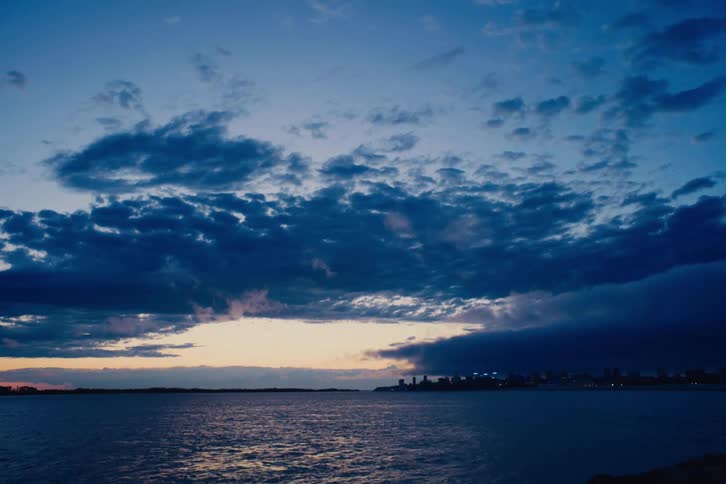 A distant city skyline is visible across the calm sea under a dramatic, dark blue sky at dusk
