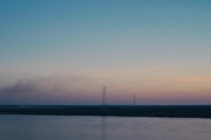 A serene wide shot of a river at dusk, with power lines silhouetted on the horizon against a pastel sky