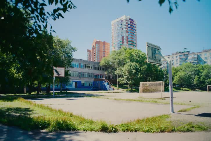 A quiet and deserted outdoor basketball court and schoolyard sits empty on a bright, sunny day with city buildings in the background