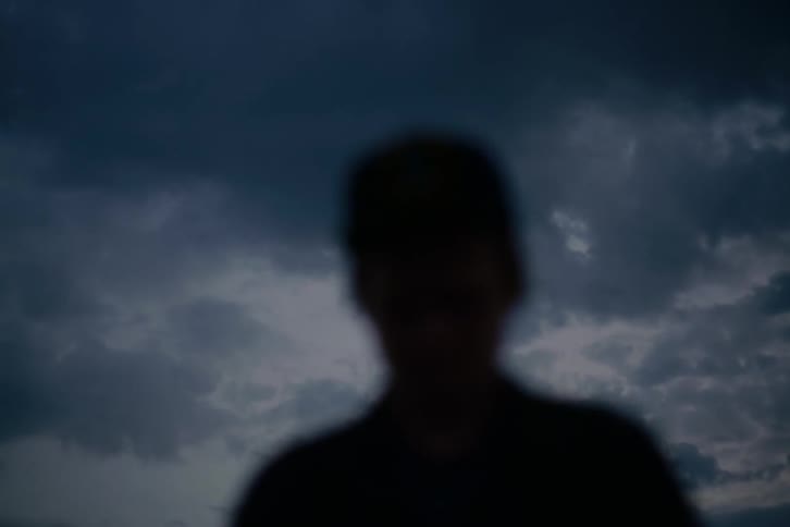 The dark, out-of-focus silhouette of a person is framed by an ominous, cloudy sky