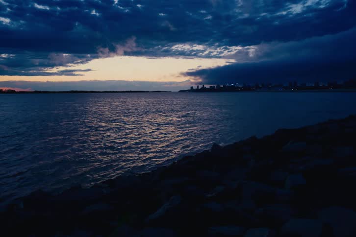 Dark, dramatic clouds gather over calm waters at dusk, with a distant city skyline glowing on the horizon