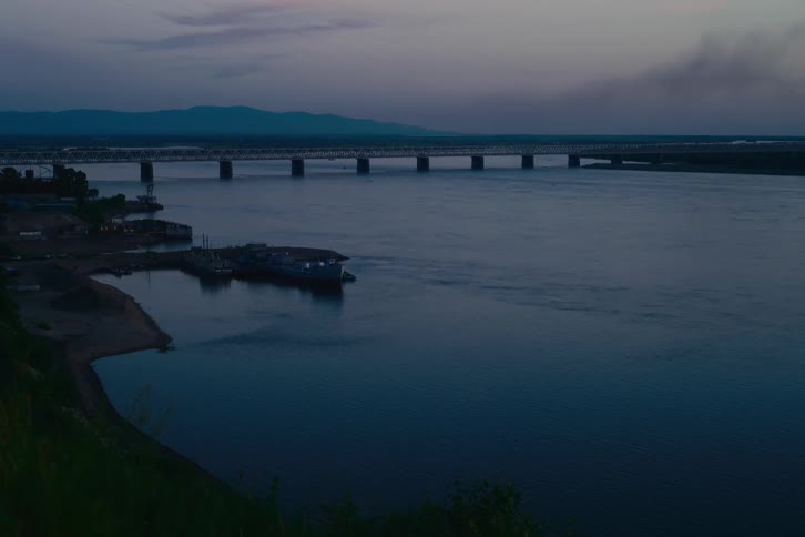 A serene wide shot of a large river and a bridge in the distance during a calm twilight