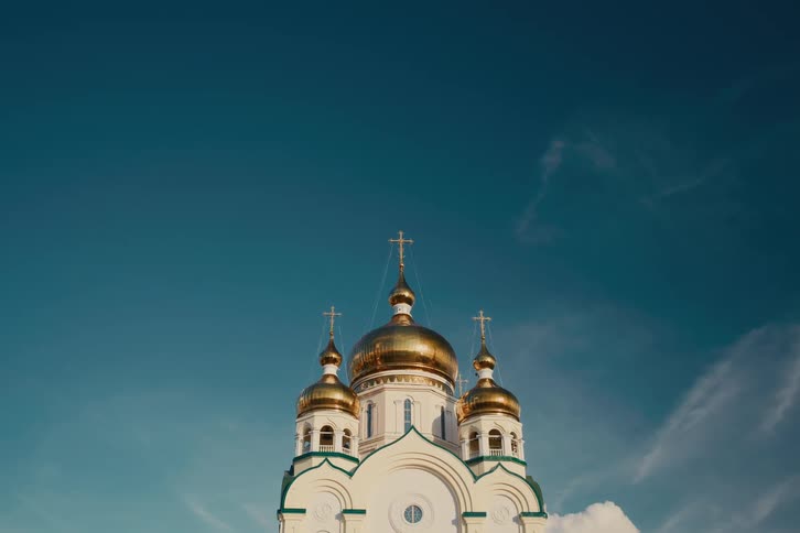 A static low-angle shot of a white Orthodox church with shining golden domes against a deep blue sky with wispy, moving clouds