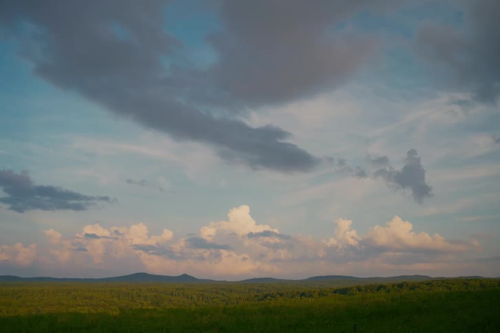 A tranquil wide shot of a rolling green landscape with forests and mountains under a beautiful cloudy sky at dusk