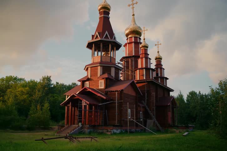 An ornate wooden Russian Orthodox church with golden domes sits in a forest clearing at sunset