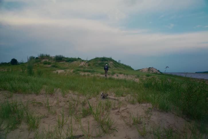A lone hiker with a backpack walks away from the camera up a grassy sand dune on an overcast day