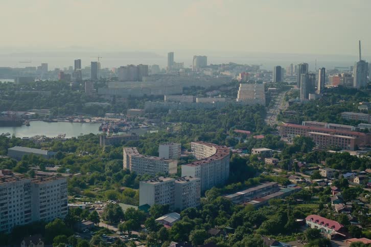 A high-angle, static daytime view of a sprawling coastal city featuring diverse architecture and a busy port under a hazy sky