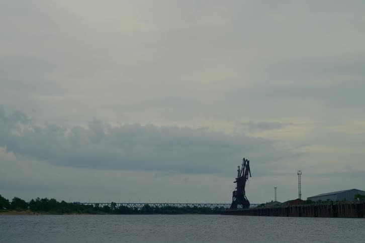 A stationary industrial crane sits on the banks of a river with a bridge in the background under an overcast sky