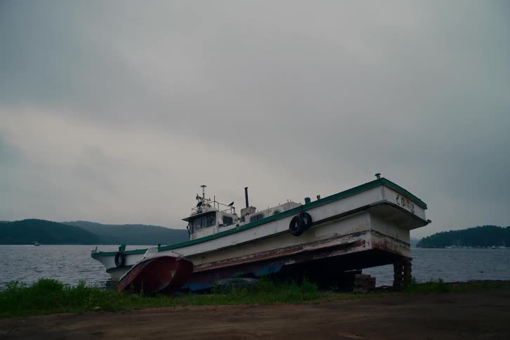 An old fishing boat and a small dinghy rest abandoned on the shore of a lake on a gloomy, overcast day