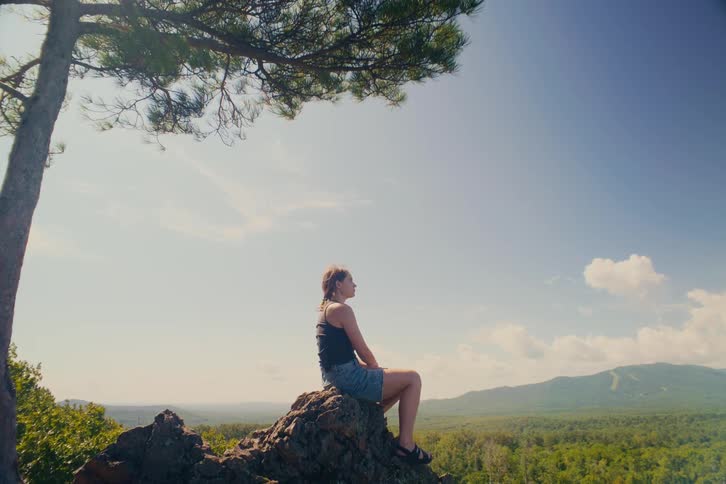 A contemplative woman sits on a rocky mountain overlook under a tree, peacefully enjoying the vast forest and mountain landscape on a sunny day