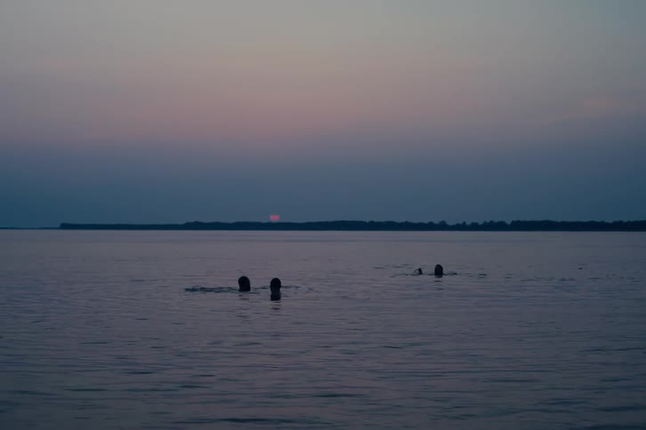 Silhouettes of three friends enjoying a peaceful swim in a vast lake at twilight as the sun sets on the horizon