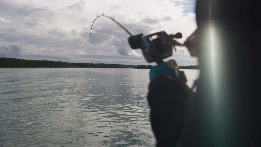An over-the-shoulder slow-motion shot of a fisherman reeling in a catch on a calm river with a bent fishing rod