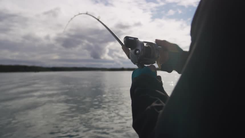 An angler on a boat reels in a catch from the water, with the bent fishing rod silhouetted against a cloudy sky in slow motion