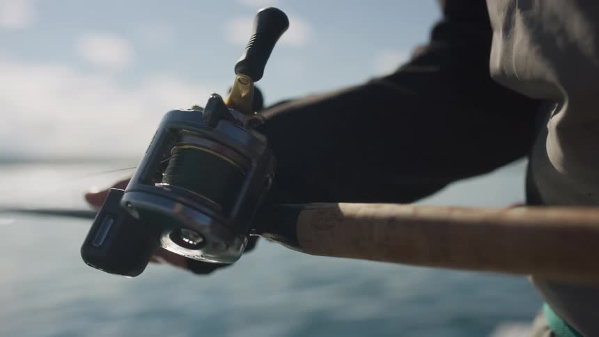 Cinematic slow-motion close-up of a fishing reel being cranked as a fisherman reels in their line over the sunny ocean
