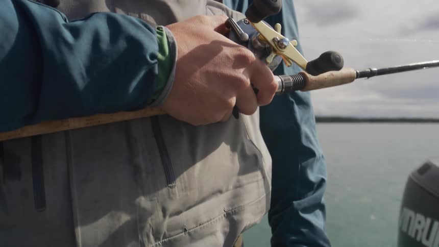 A fisherman in full waders stands on a boat and holds his fishing rod and reel while waiting for a bite