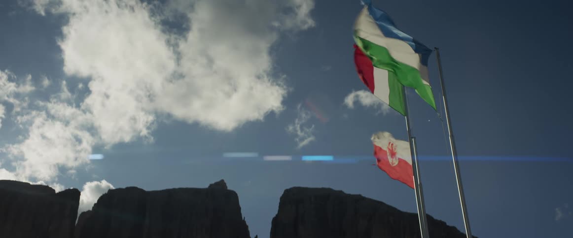 The flags of Italy, South Tyrol, and the Ladin people wave in slow motion with a silhouetted mountain range in the background