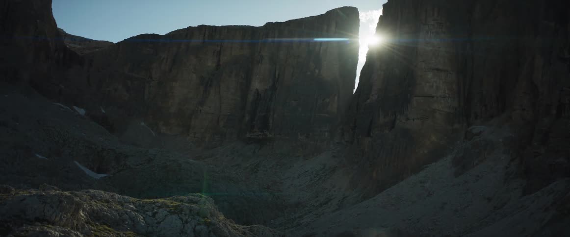 The sun peeks through a crevice in a vast rocky mountain landscape, creating a dramatic lens flare as the camera slowly pans
