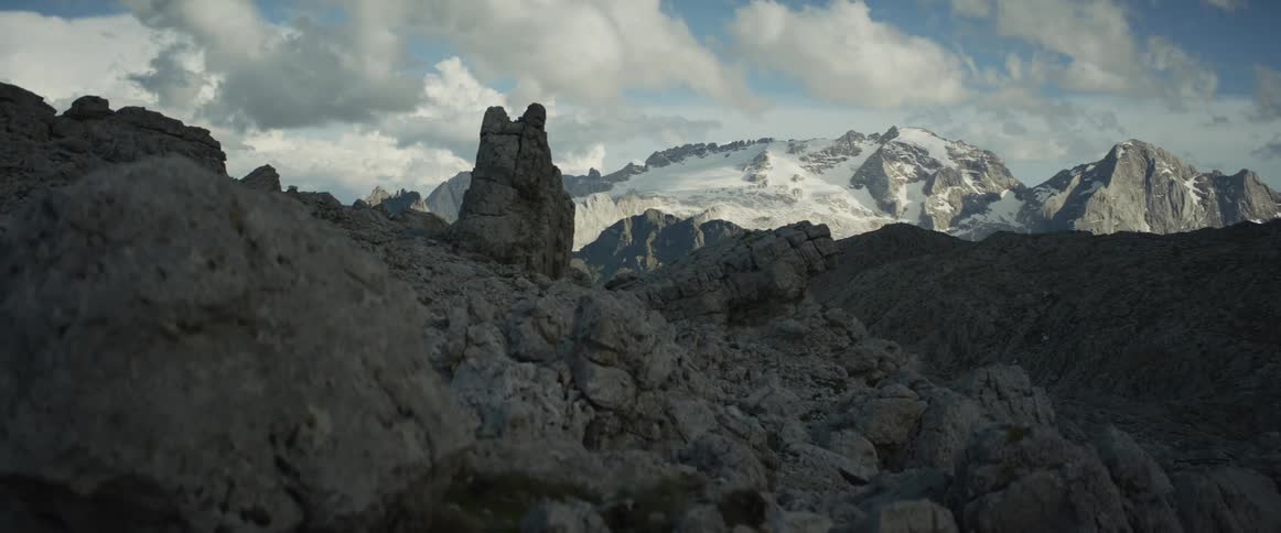 A rocky, shadowed foreground gives way to a sunlit, snow-capped mountain range under a cloudy sky