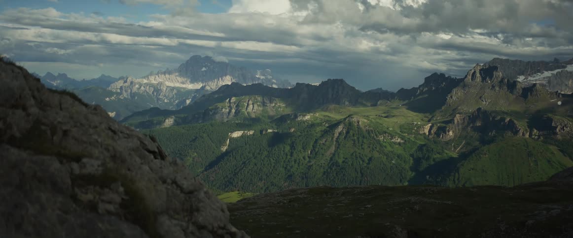 Sunlight breaks through dramatic clouds, casting moving shadows across a vast and majestic mountain valley