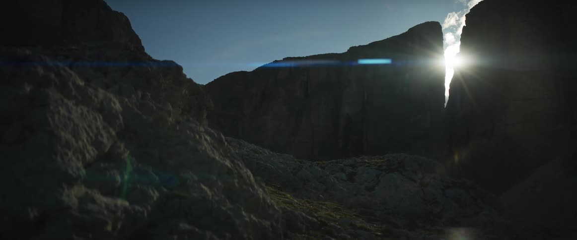 The sun shines brightly from between two silhouetted mountain peaks, creating an epic lens flare over the rocky landscape