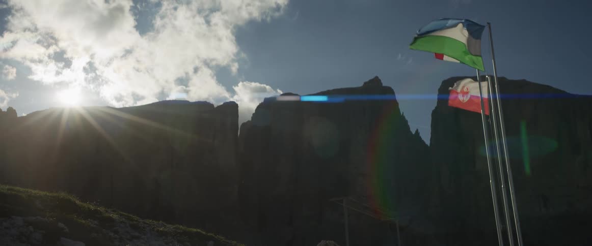 Cinematic slow motion shot of Ladin and South Tyrol flags waving against the stunning silhouetted Sella massif with a beautiful lens flare