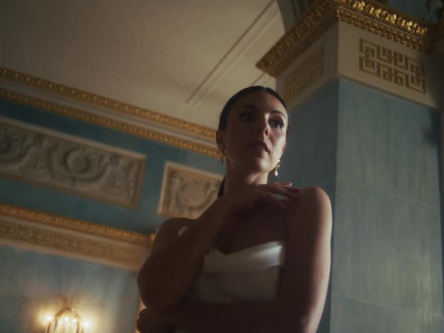 A beautiful, contemplative woman in a white dress stands in a lavish, ornate room with blue and gold walls