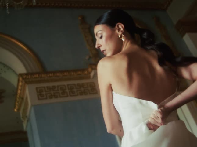 In an opulent, historic room, an elegant woman zips up her flowing white strapless gown in cinematic slow motion