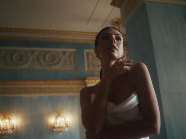 From a low angle, an elegant woman in a white dress poses thoughtfully inside a luxurious blue and gold room