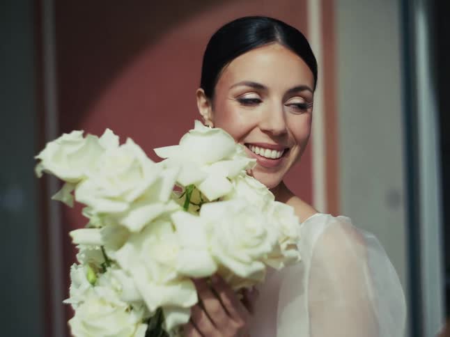 A beautiful, joyful bride with dark hair pulled back smiles brightly while holding a lovely bouquet of white roses