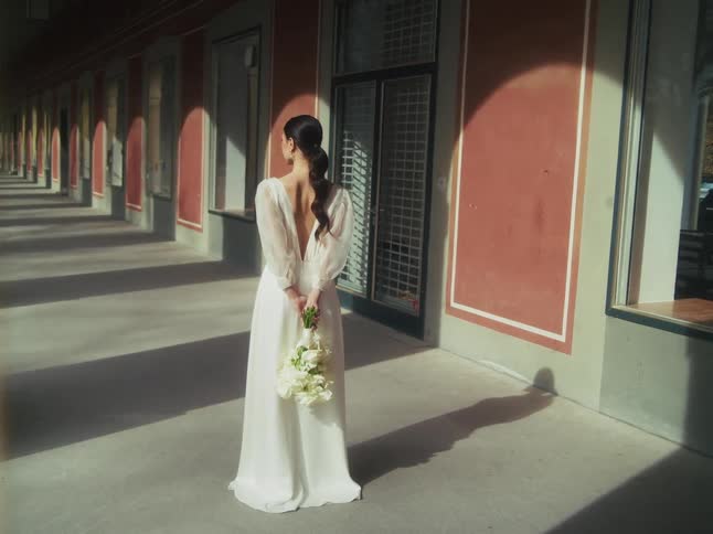 An elegant bride in a white gown holds a bouquet behind her back while standing in a sun-drenched architectural colonnade