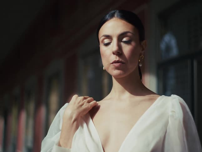 A thoughtful and elegant woman in a white dress poses in the dramatic sunlight of an outdoor corridor