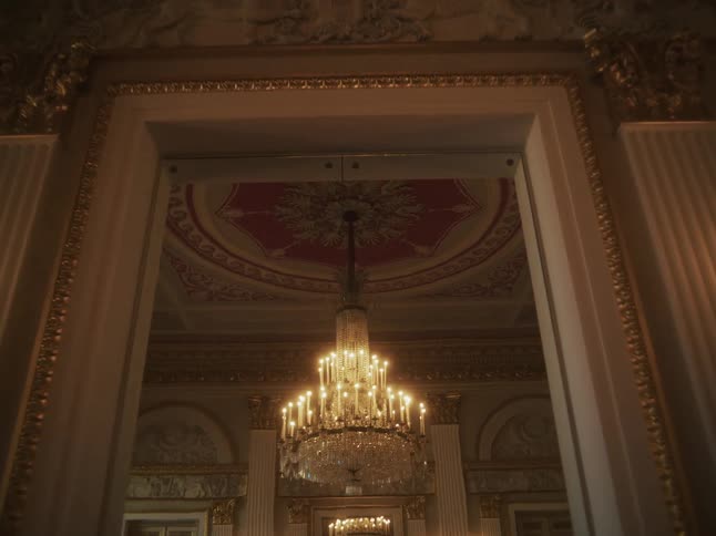 A slow dolly shot reveals an ornate crystal chandelier hanging from a lavishly decorated ceiling in a grand historical ballroom