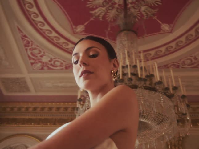 An elegant woman wrapped in a white towel smiles flirtatiously at the camera while standing in a luxurious, ornate room