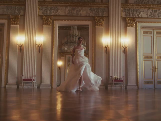 An elegant bride in a flowing white wedding dress walks gracefully towards the camera in a grand, empty ballroom