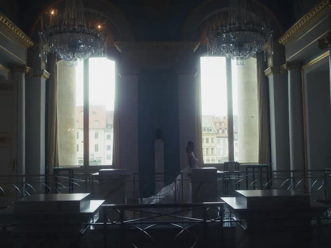 An elegant woman in a white dress walks through a dimly lit, opulent hall with large windows and crystal chandeliers