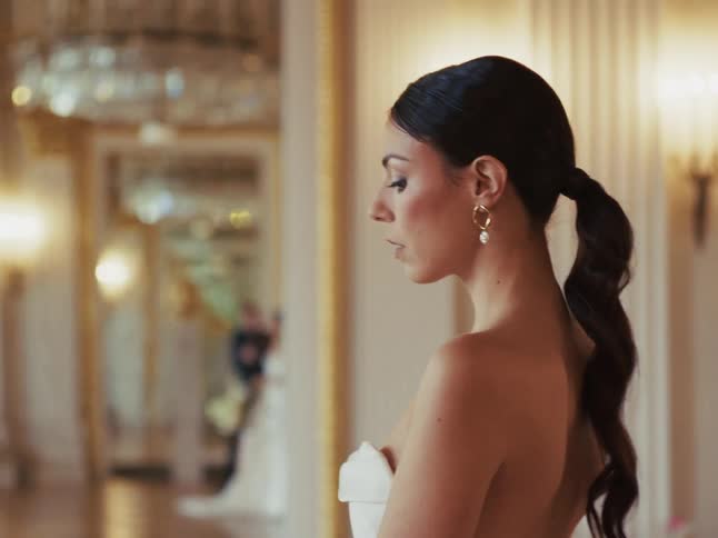 An elegant bride in a beautiful, softly lit room thoughtfully smells her white rose bouquet