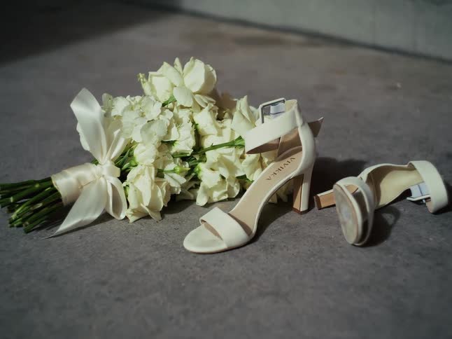 Still life shot of a bride's white high-heeled sandals and a white flower bouquet on a concrete floor
