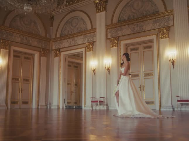 A gorgeous bride in a long white dress stands thoughtfully in an opulent, empty ballroom