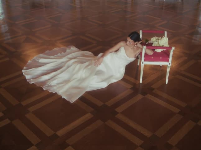 A beautiful bride in a flowing white gown lies on an ornate wooden floor in a grand room, posing elegantly next to a chair with her bouquet