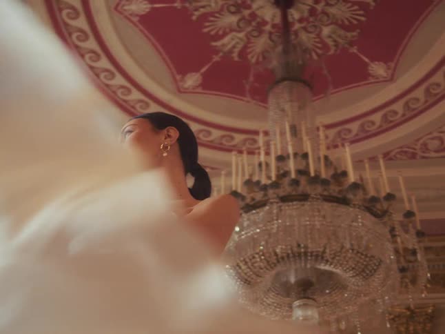 Low-angle slow motion shot of a beautiful woman in a white gown posing in a magnificent ballroom with an ornate ceiling