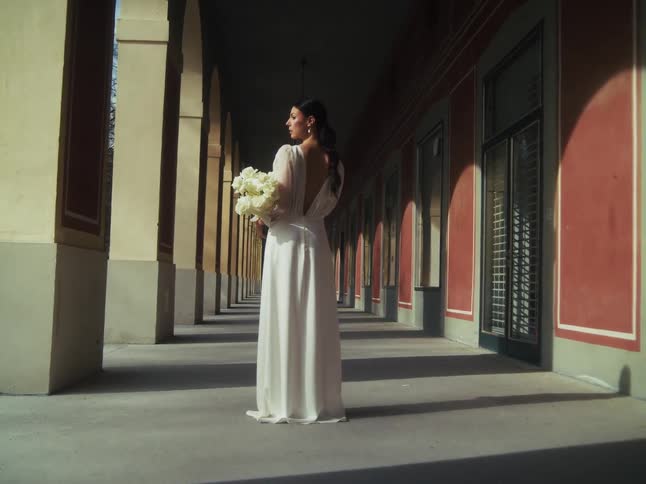 An elegant bride in a white wedding gown holds a bouquet while posing in a beautiful, sun-drenched colonnade