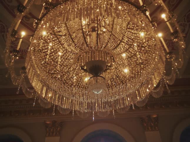 A slow tilt-up shot reveals the intricate details of a massive, glowing crystal chandelier in a grand, historic hall