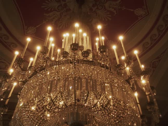 A grand, multi-tiered crystal chandelier with glowing candle-like bulbs hangs from a decorative red ceiling in a luxurious hall