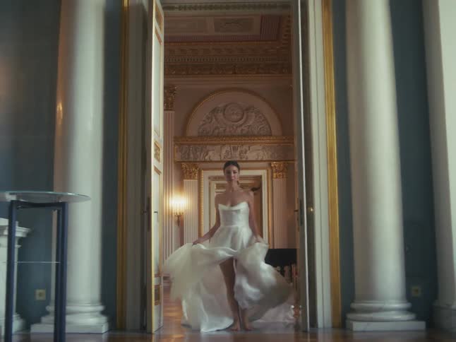 A beautiful bride in a flowing white gown walks barefoot towards the camera in slow motion through a luxurious and ornate palace hall