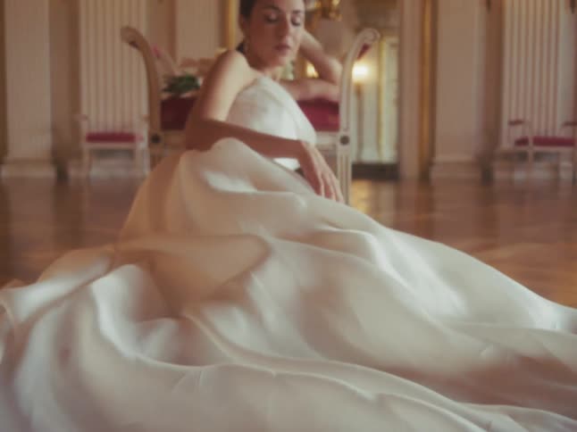 Rack focus from the fabric of a white wedding dress to a beautiful bride sitting gracefully in a magnificent ballroom
