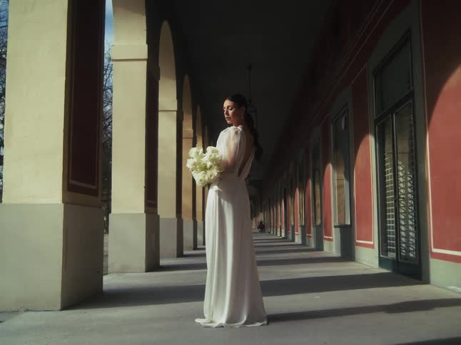 A beautiful bride in a flowing white gown stands gracefully with a bouquet in a long, sunlit colonnade